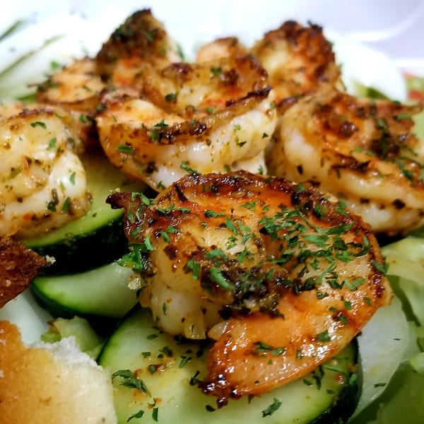 Close-up of grilled shrimp seasoned with herbs, served over sliced cucumbers and lettuce.